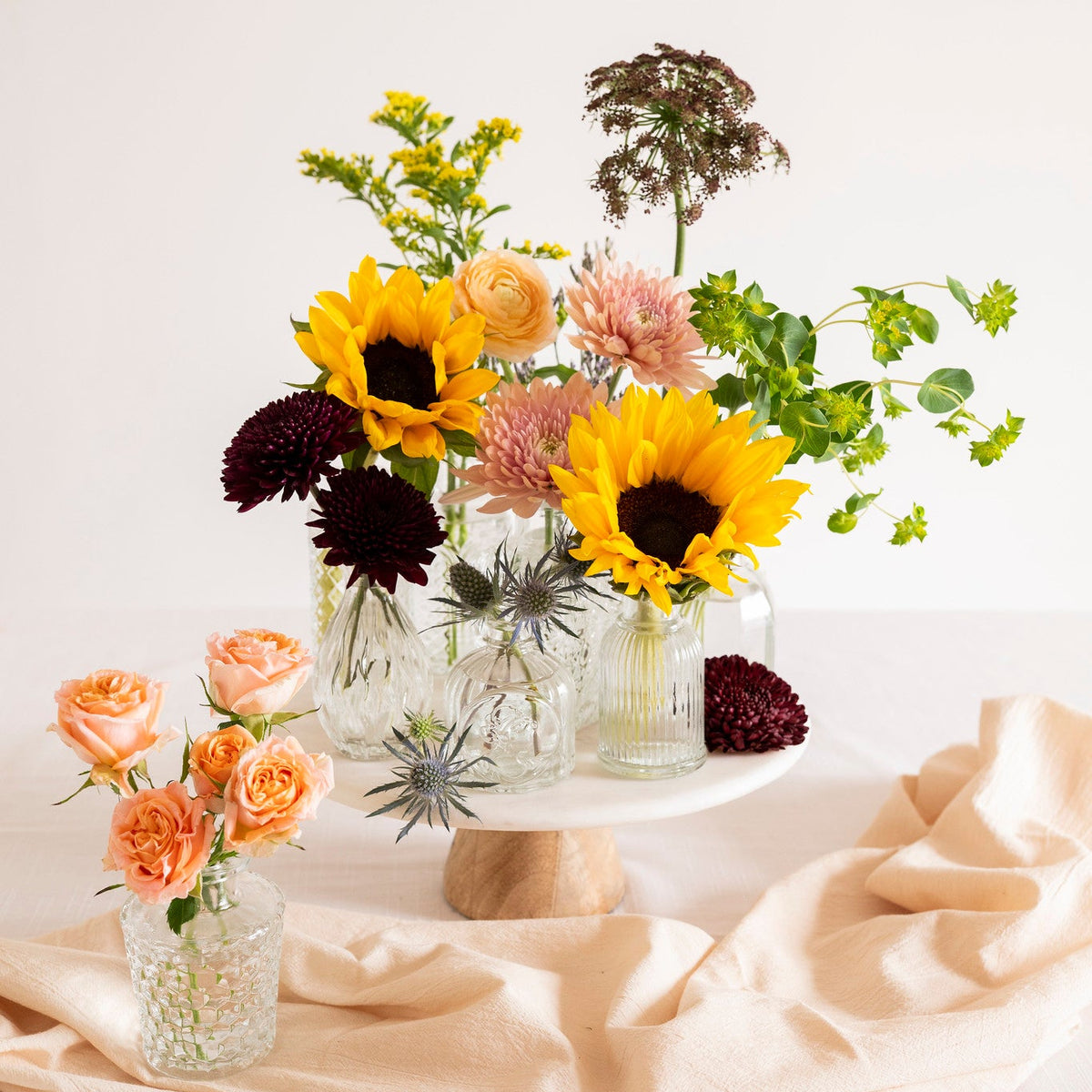 A floral arrangement in clear glass jars and vases on a white pedestal stand, styled with peach roses, yellow sunflowers, pink blooms, and green foliage on a draped cream fabric surface against a white background.