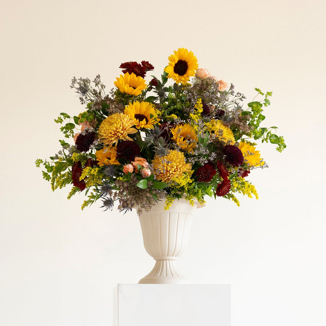 A floral arrangement featuring yellow sunflowers, orange chrysanthemums, burgundy dahlias, and green foliage, presented in a white classical-style urn on a white pedestal against a warm off-white background.