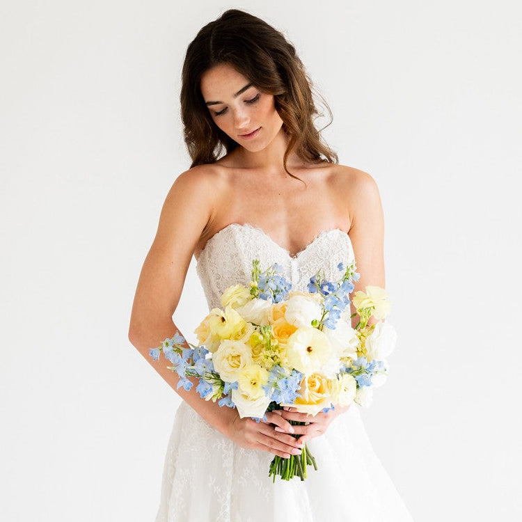 A woman wearing a strapless white wedding gown with a long train, holding a bouquet of yellow and light blue flowers, photographed against a plain white studio background.
