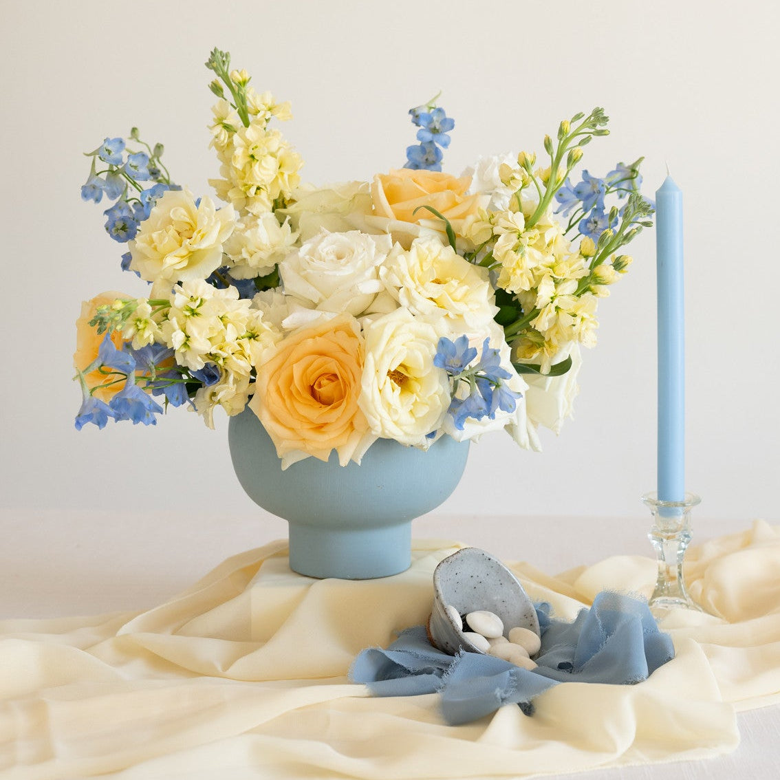 A pastel floral arrangement in a light blue vase on draped cream fabric, with a matching blue candle in a glass holder and a small blue fabric accessory in front.