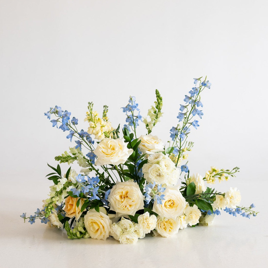 A small floral arrangement featuring cream and pale yellow roses with blue delphiniums and green foliage, displayed on a clean white surface against a white background.