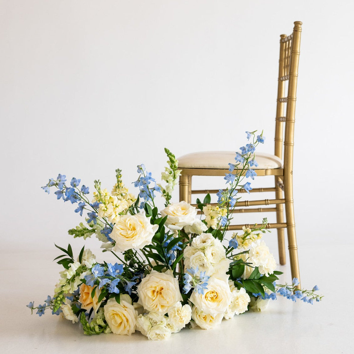 A gold wooden chair with a cream upholstered seat is positioned beside a large floral arrangement of white and pale yellow roses with blue flowers and green foliage on a clean white studio background.