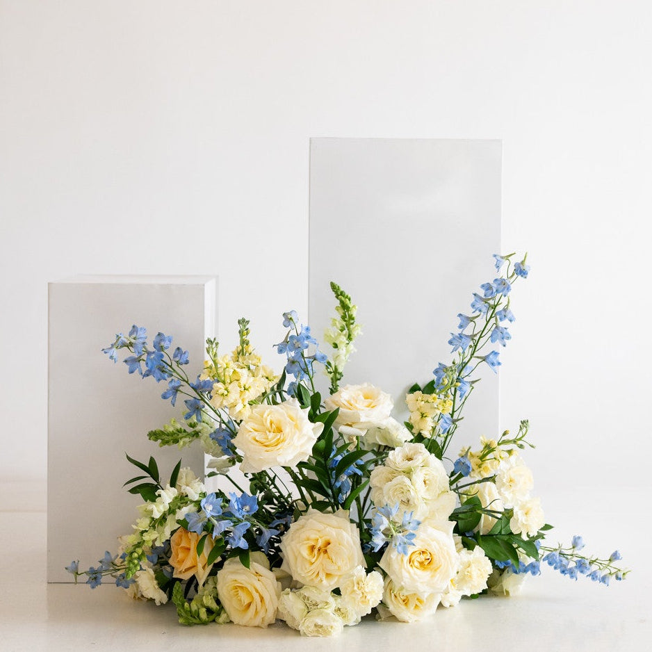 A floral arrangement with pale yellow roses, white flowers, and blue blossoms placed in front of two white rectangular cards on a clean white surface against a white background.