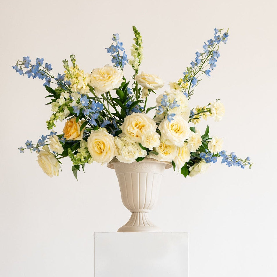A floral arrangement featuring pale yellow roses and blue delphiniums in a white classical urn vase, displayed on a white rectangular pedestal against a clean white background.