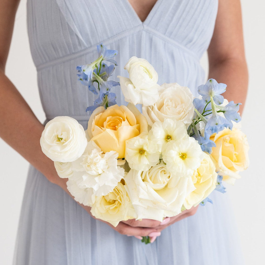 A woman wearing a light blue pleated tulle dress holds a bouquet of white and pale yellow flowers with small blue accents against a plain white background.