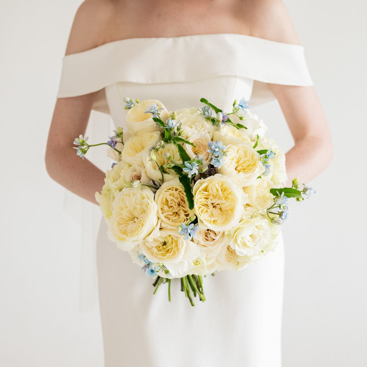 A bride in an off-the-shoulder white wedding dress holding a large bouquet of cream and pale yellow roses with small blue flowers, photographed against a clean white background.