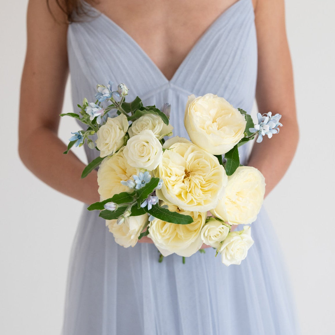 A woman wearing a light blue sleeveless tulle dress holds a bouquet of pale yellow flowers with green leaves and small light blue blossoms against a plain light background.