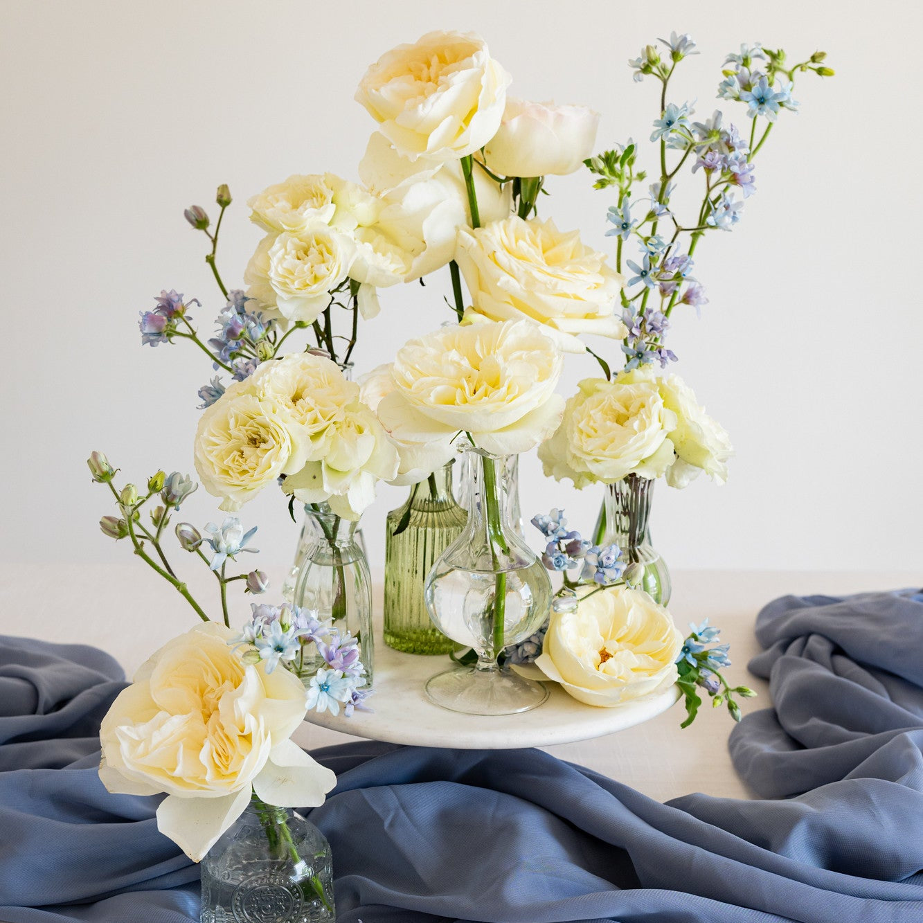 A floral centerpiece arrangement of pale yellow roses, white blooms, and light blue flowers in clear glass vases and a small green glass bottle, styled on a white surface with draped slate-blue fabric against a warm off-white background.