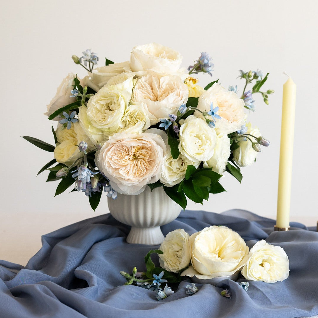 A floral arrangement of cream and pale peach roses with green foliage in a white ribbed vase, styled on draped slate blue fabric with two lit cream taper candles against a soft off-white background.