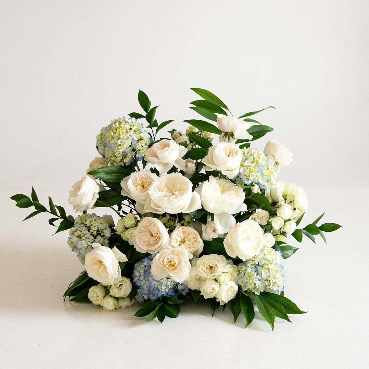 A lush floral arrangement featuring cream and white roses, light blue hydrangeas, and green foliage, presented on a clean white studio background.