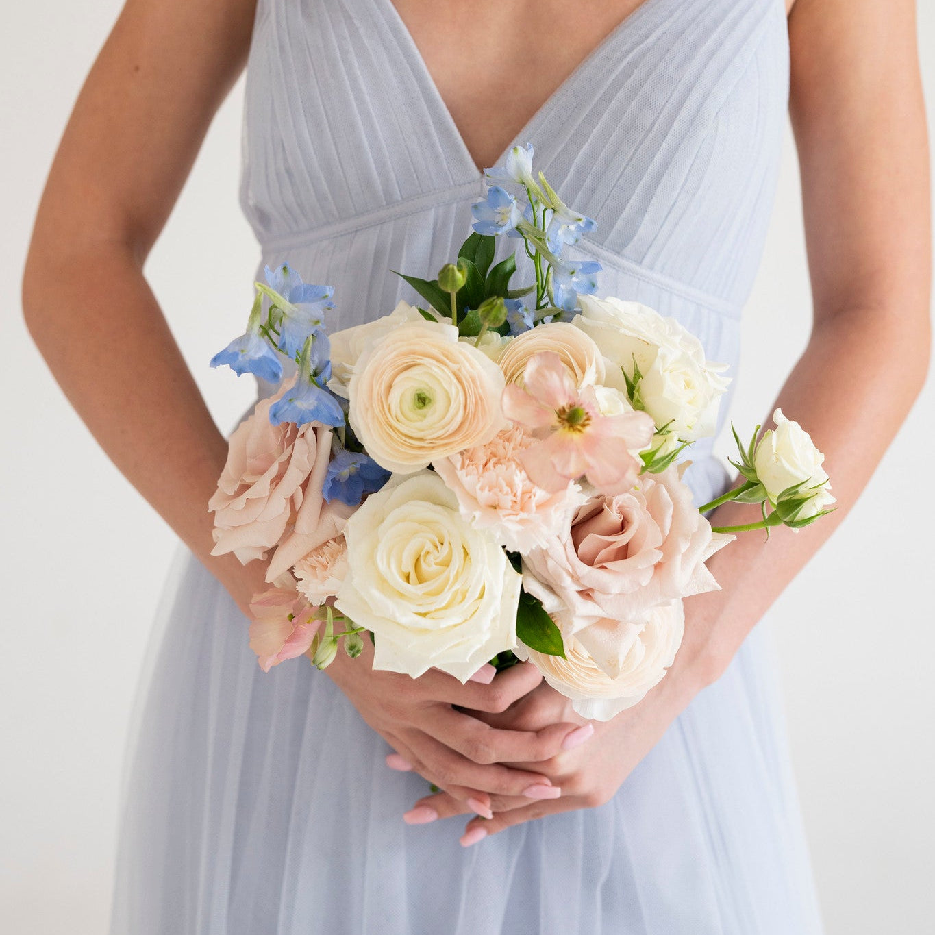 A woman wearing a light blue pleated spaghetti-strap dress holds a bridal bouquet of blush pink, cream, and light blue flowers against a plain white background.