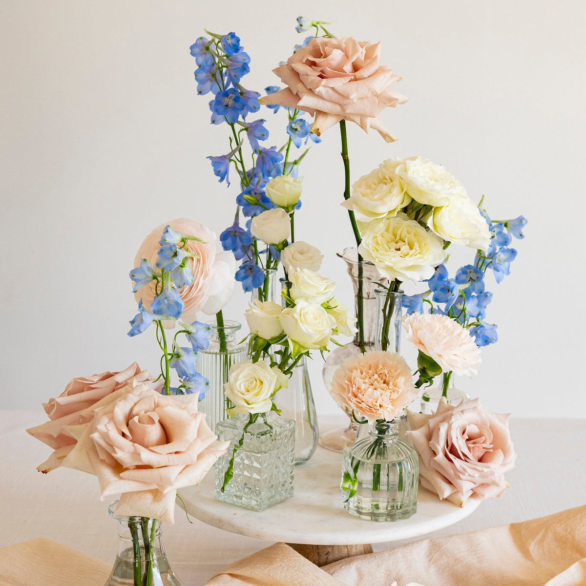 A styled floral arrangement of blush pink, cream, and blue flowers in clear glass vases, displayed on a white pedestal with draped peach fabric against a soft cream background.