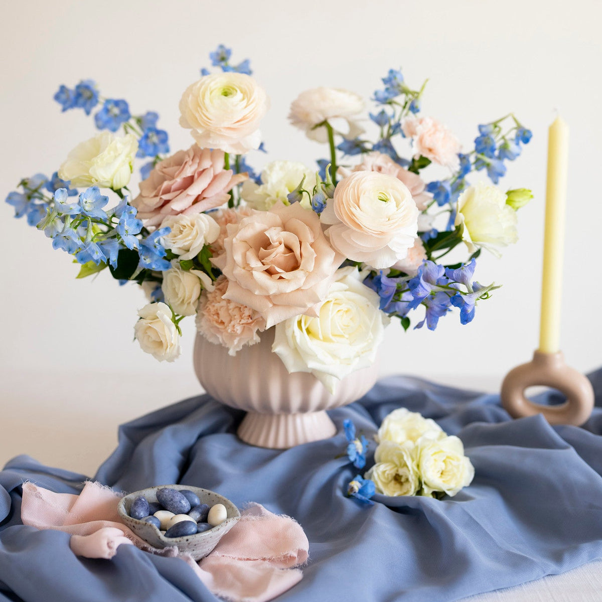 A floral arrangement in a ribbed white ceramic vase, featuring blush pink and cream roses with blue delphiniums, styled on a draped slate blue fabric with a pale yellow candle and small decorative accents.