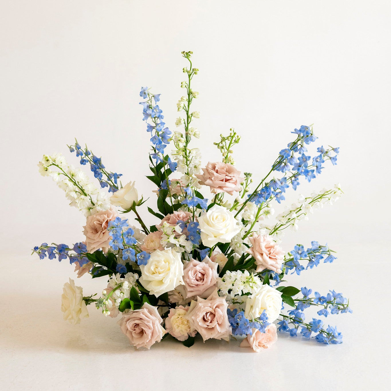 A delicate floral arrangement featuring light pink and cream roses with blue delphiniums and green foliage, set against a clean white background.