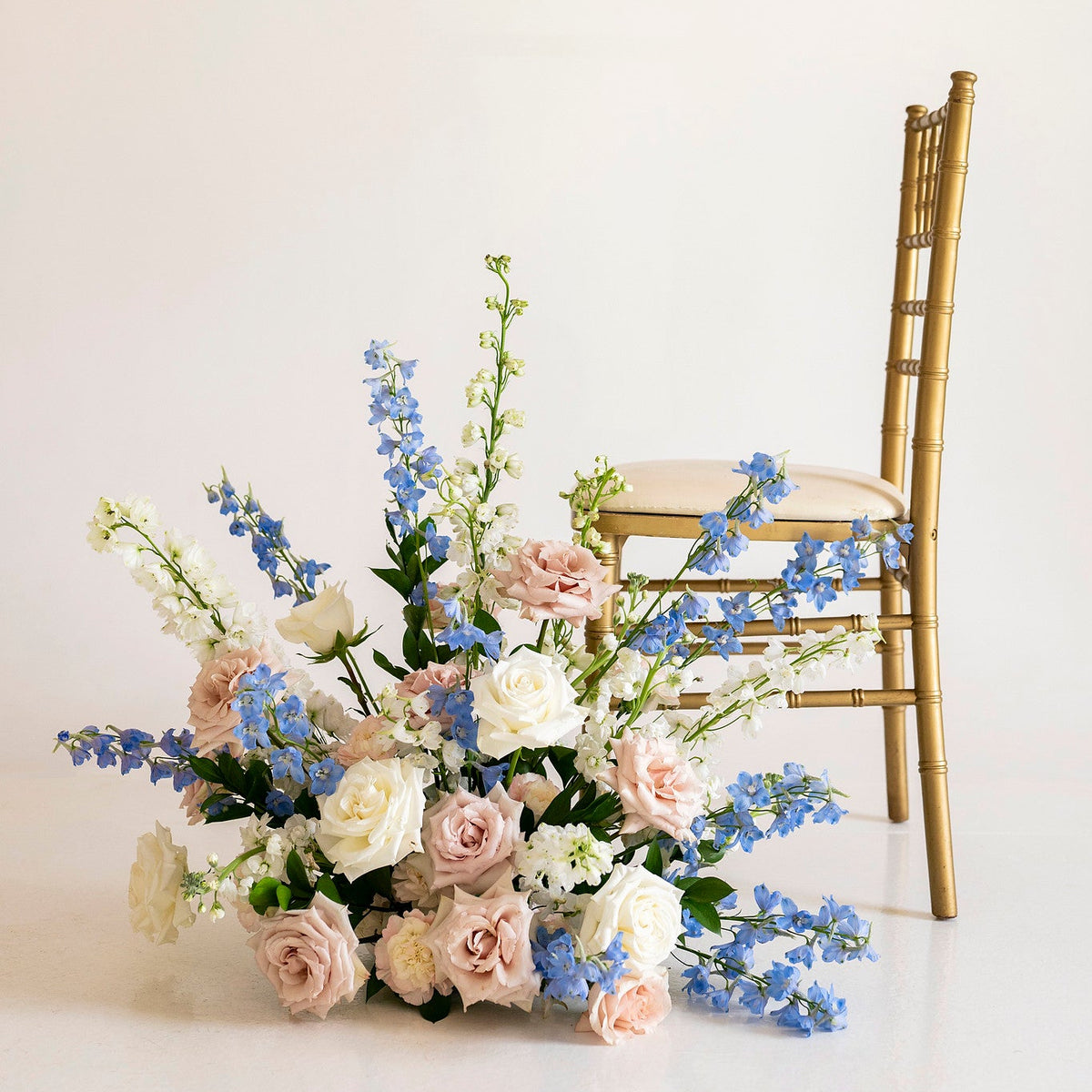 A floral arrangement of pink and white roses with blue delphiniums and green foliage, styled next to a gold Chiavari chair against a clean white background.