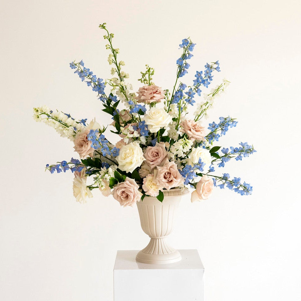 A floral arrangement in a white urn-shaped vase displayed on a white pedestal against a plain off-white background.