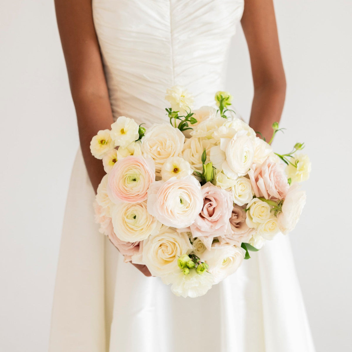 A woman wearing a white wedding dress holds a large bouquet of pale pink, cream, and white flowers with green leaves, photographed against a soft light background.