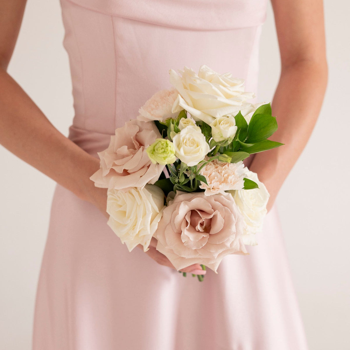 A close-up of a person wearing a light pink satin dress and holding a bouquet of pale pink and white roses with green leaves, set against a soft light background.