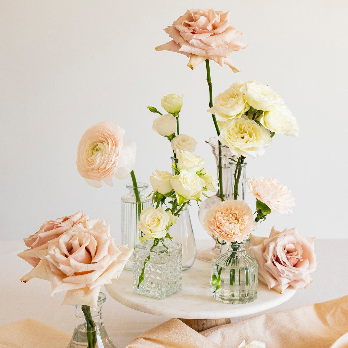 A styled floral arrangement of pale blush, cream, and peach roses and ranunculus in clear glass vases, displayed on a white pedestal with draped peach fabric against a soft off-white background.