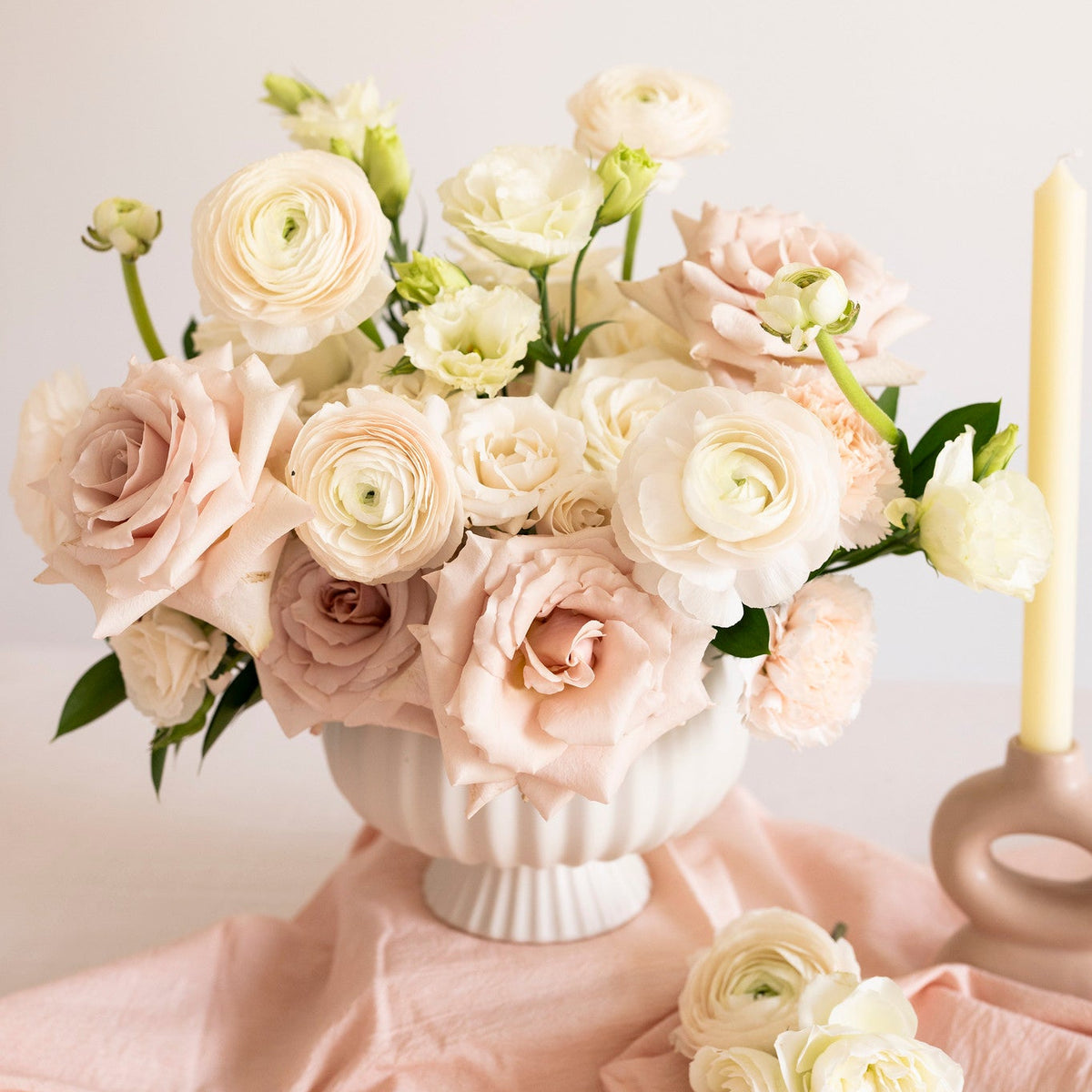 A bouquet of blush pink and cream ranunculus flowers arranged in a white ribbed ceramic vase, set on draped blush fabric with a matching flower at the base and a lit candle holder beside it.