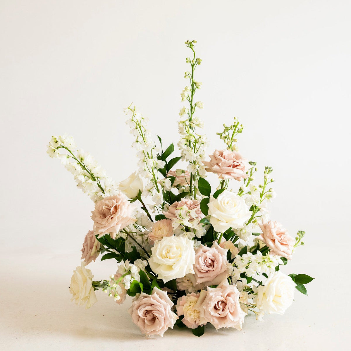 A delicate floral arrangement featuring blush pink and white roses, white delphiniums, and green foliage, centered against a clean, off-white background.