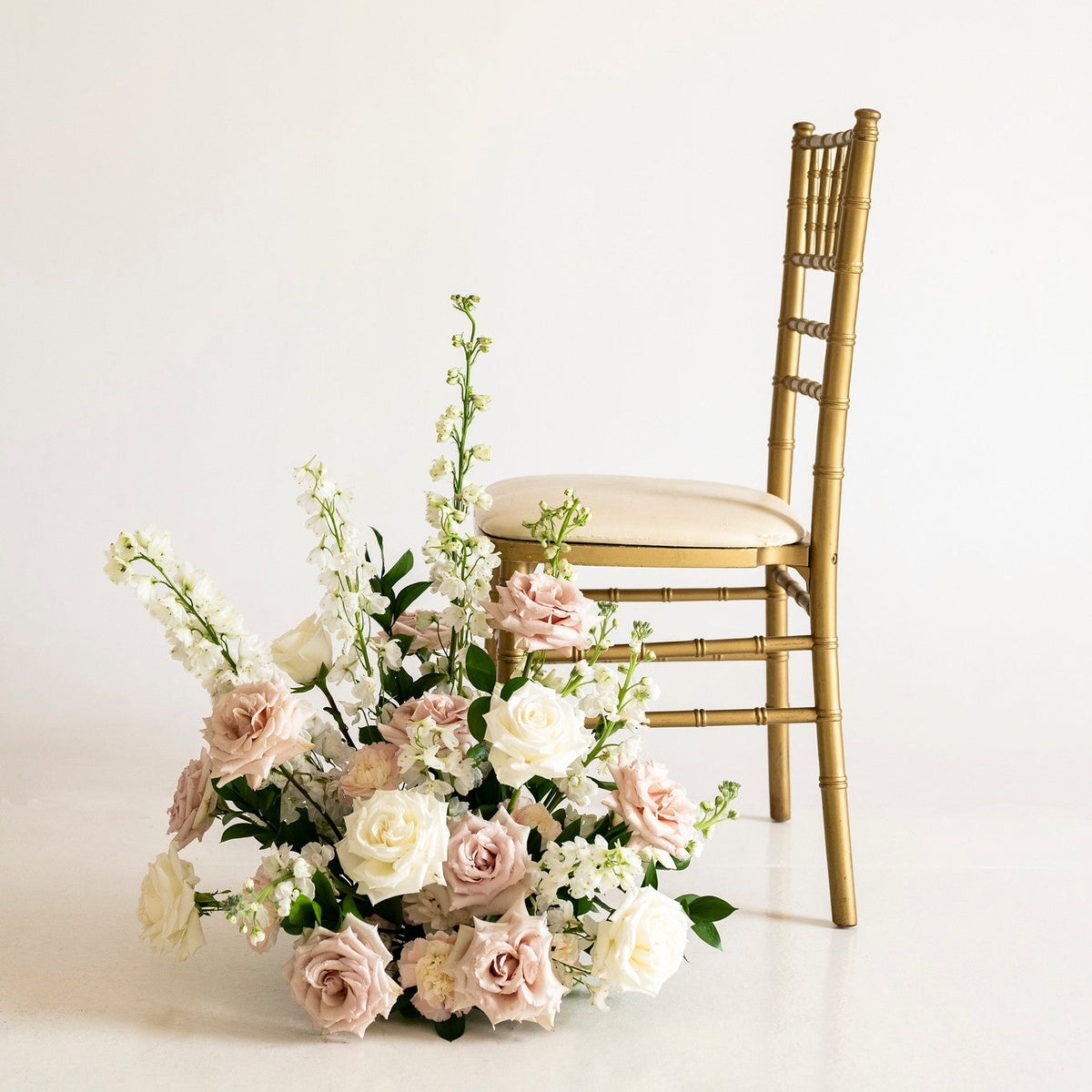 A gold-finished wooden chair with a cream upholstered seat is positioned beside a large floral arrangement of blush pink, cream, and white roses with green foliage on a clean white studio background.