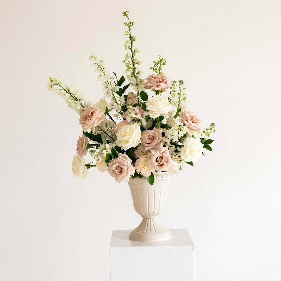A floral arrangement in a white urn-shaped vase displayed on a white pedestal against a plain off-white background.