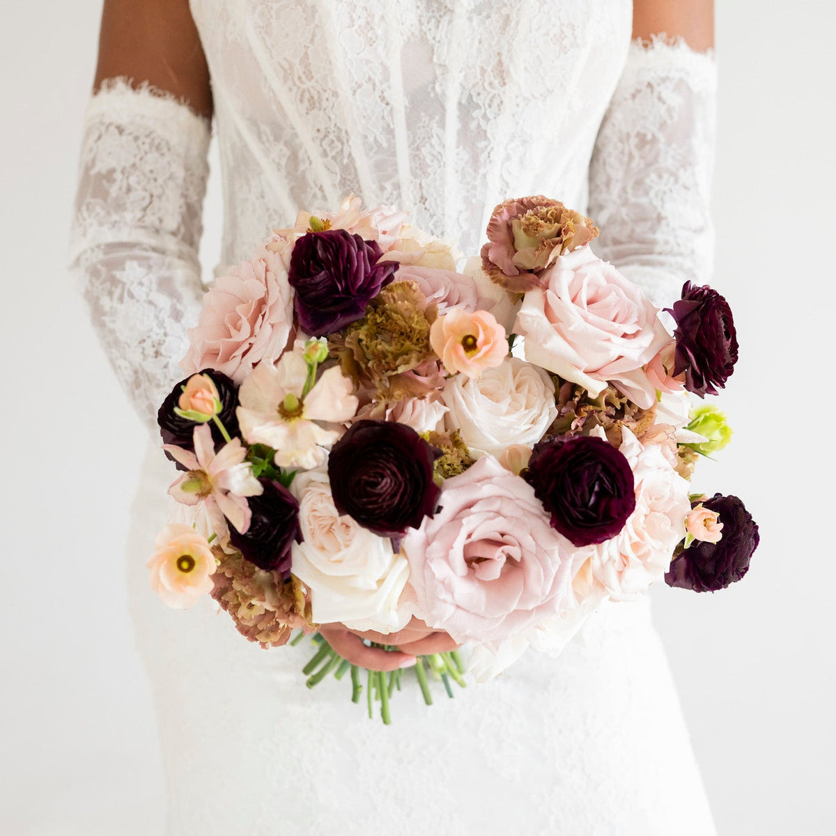 A bride in a white lace wedding dress and matching lace gloves holds a large bouquet of blush pink, deep burgundy, and cream flowers against a clean white background.