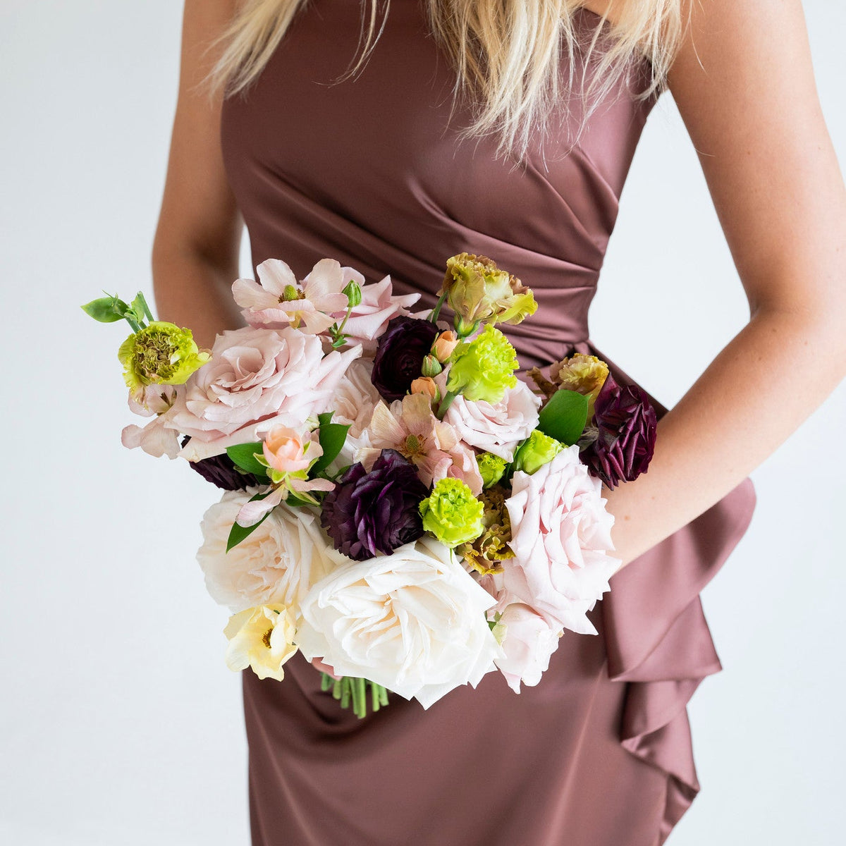 A woman wearing a satin-like mauve-brown strapless dress holds a bouquet of pale pink, white, and deep purple flowers against a clean white background.