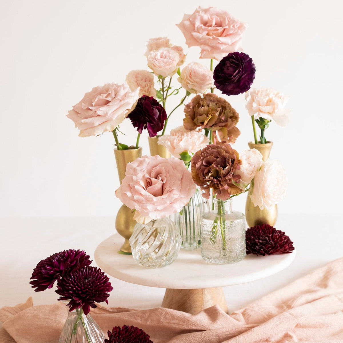 A styled floral arrangement featuring light pink and deep burgundy roses in clear glass vases and gold vases, displayed on a white pedestal cake stand with a peach fabric drape underneath.