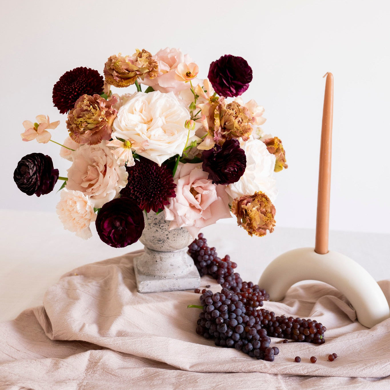 A styled still life featuring a bouquet of dark burgundy, blush pink, and tan dried-style flowers in a stone vase, set on a beige cloth with dark grapes and a cream candle holder with a single lit candle against a warm off-white background.