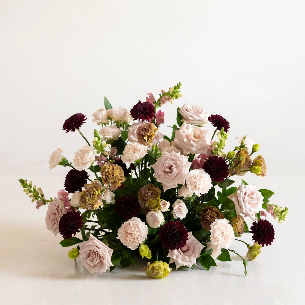A mixed floral arrangement featuring blush pink roses, deep burgundy chrysanthemums, and cream-colored flowers with green foliage, set against a plain off-white background.
