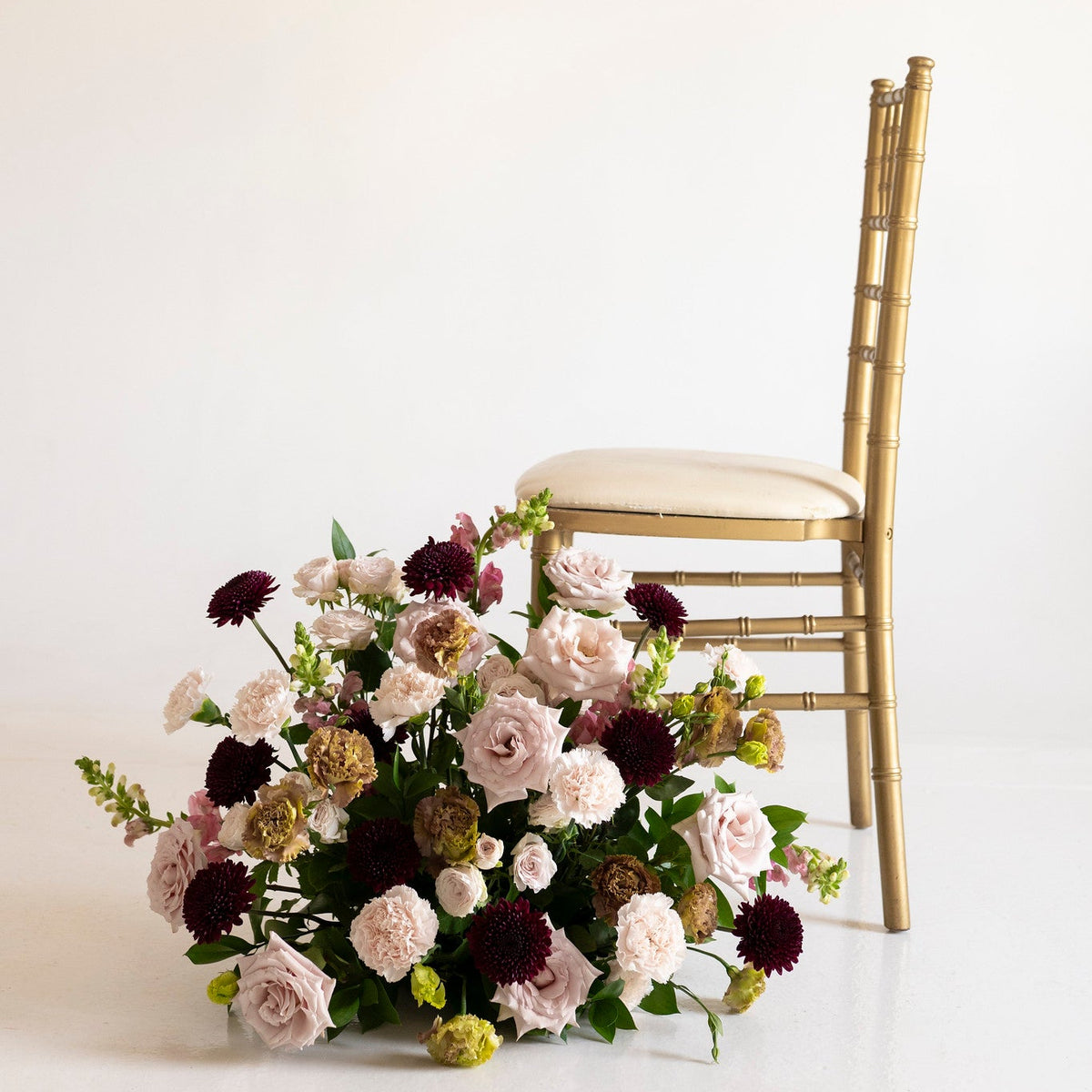 A large floral arrangement with blush pink, burgundy, cream, and olive-gold flowers sits on the floor beside a gold bamboo-style chair with a cream seat cushion against a plain off-white studio background.
