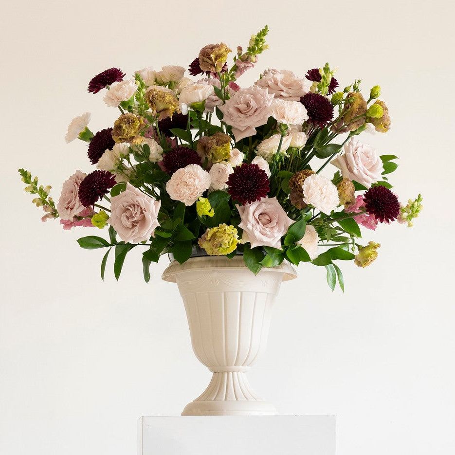A floral arrangement in a white urn-shaped vase displayed on a white pedestal against a plain white background.