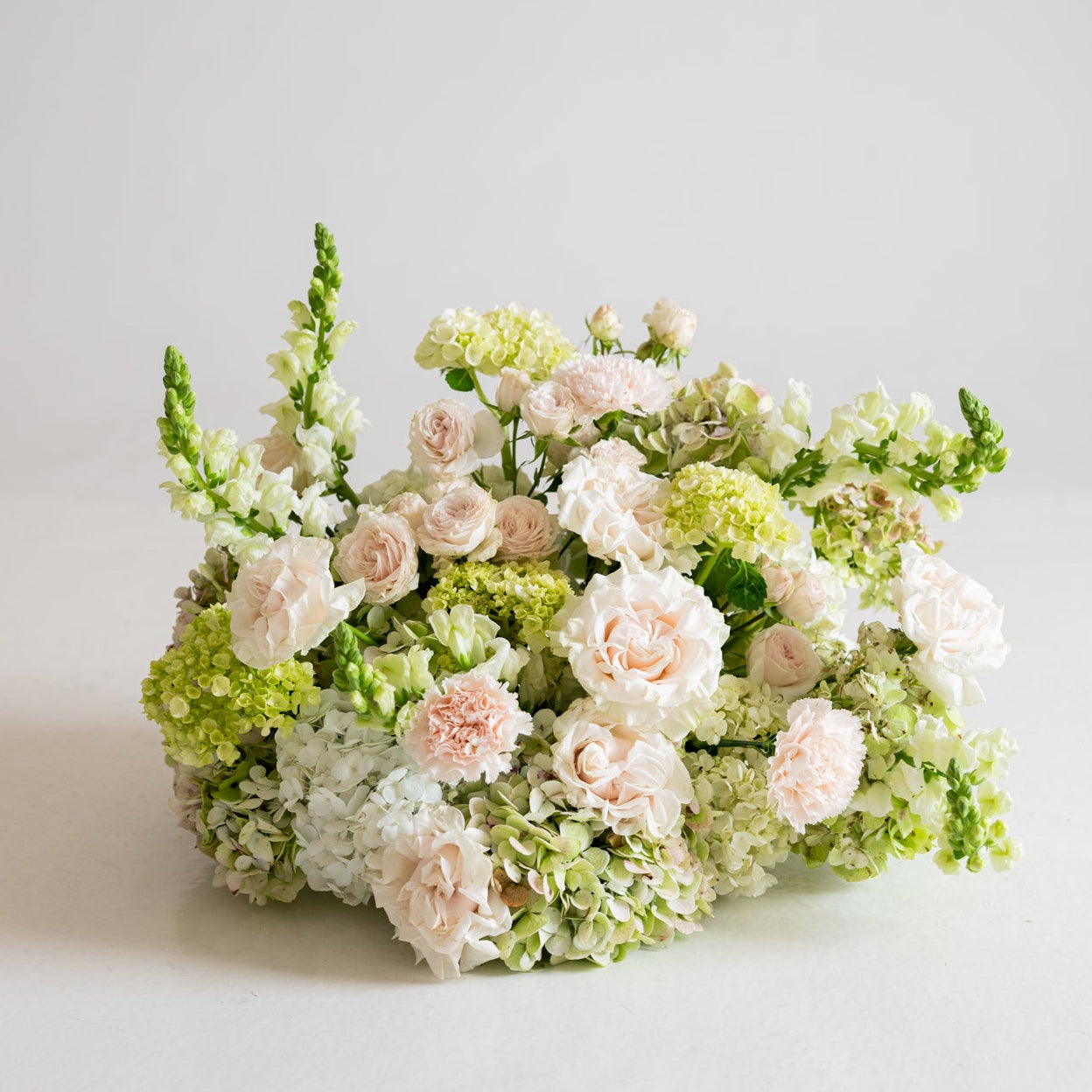 A round floral arrangement featuring pale pink roses, white flowers, and light green hydrangeas and filler stems, photographed on a clean white studio background.