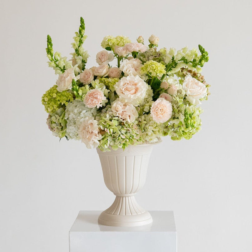 A floral arrangement of pale pink and cream flowers with green foliage, displayed in a white urn-shaped vase on a white pedestal against a light gray studio background.