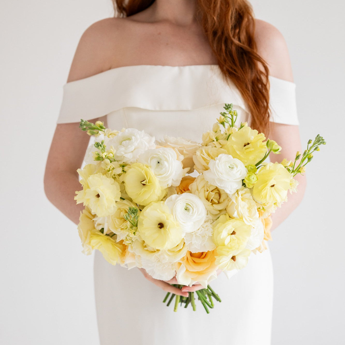 A woman wearing an off-the-shoulder white dress holds a large bouquet of yellow and white flowers, including ranunculus, against a plain white background.