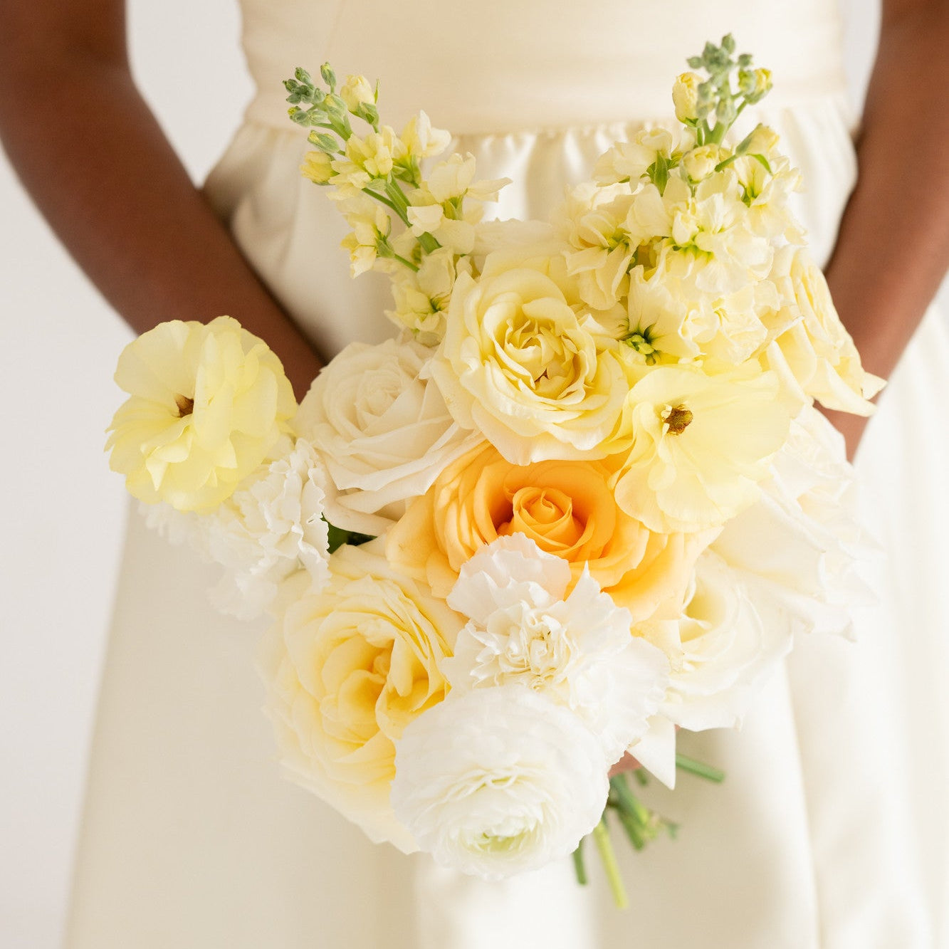 A close-up of a bouquet of yellow and white flowers held by a person wearing a cream satin dress against a soft white background.