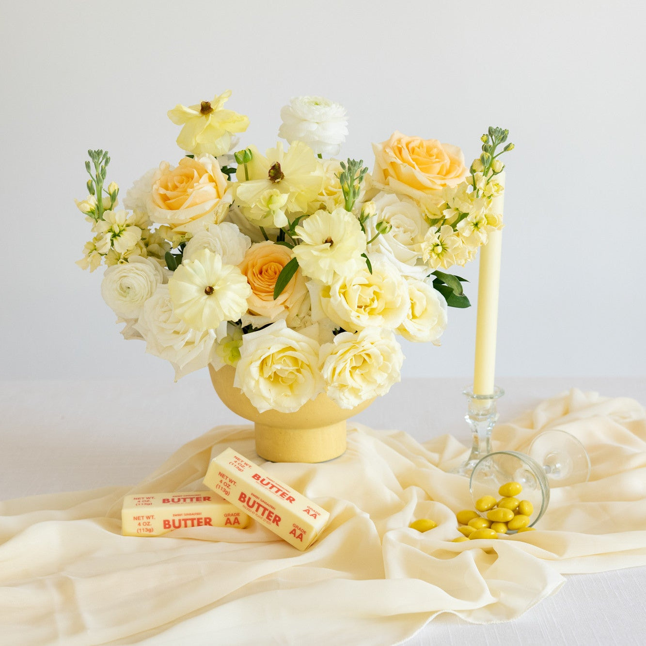 A styled floral arrangement in a yellow vase on a draped cream fabric surface, with butter sticks, a candle, and small yellow candies nearby against a soft light background.