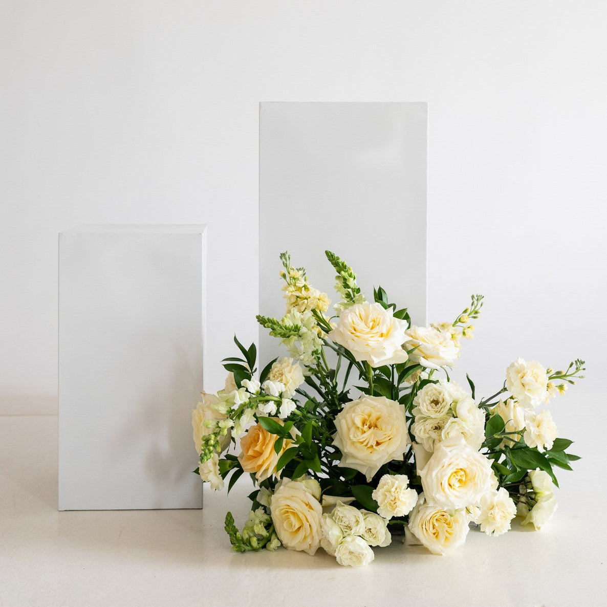 A bouquet of white and pale yellow roses with green foliage arranged in front of two minimalist white rectangular display blocks on a clean white surface against a white wall.