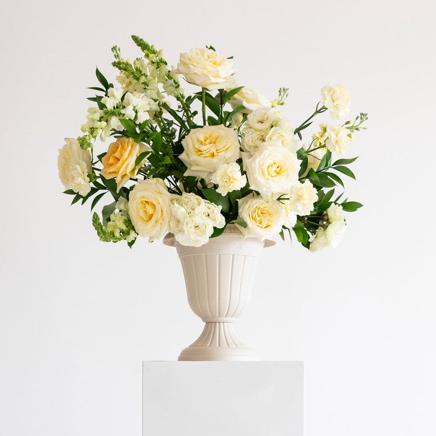 A bouquet of white and pale yellow roses with green foliage arranged in a white urn-shaped vase, displayed on a white rectangular pedestal against a clean white background.