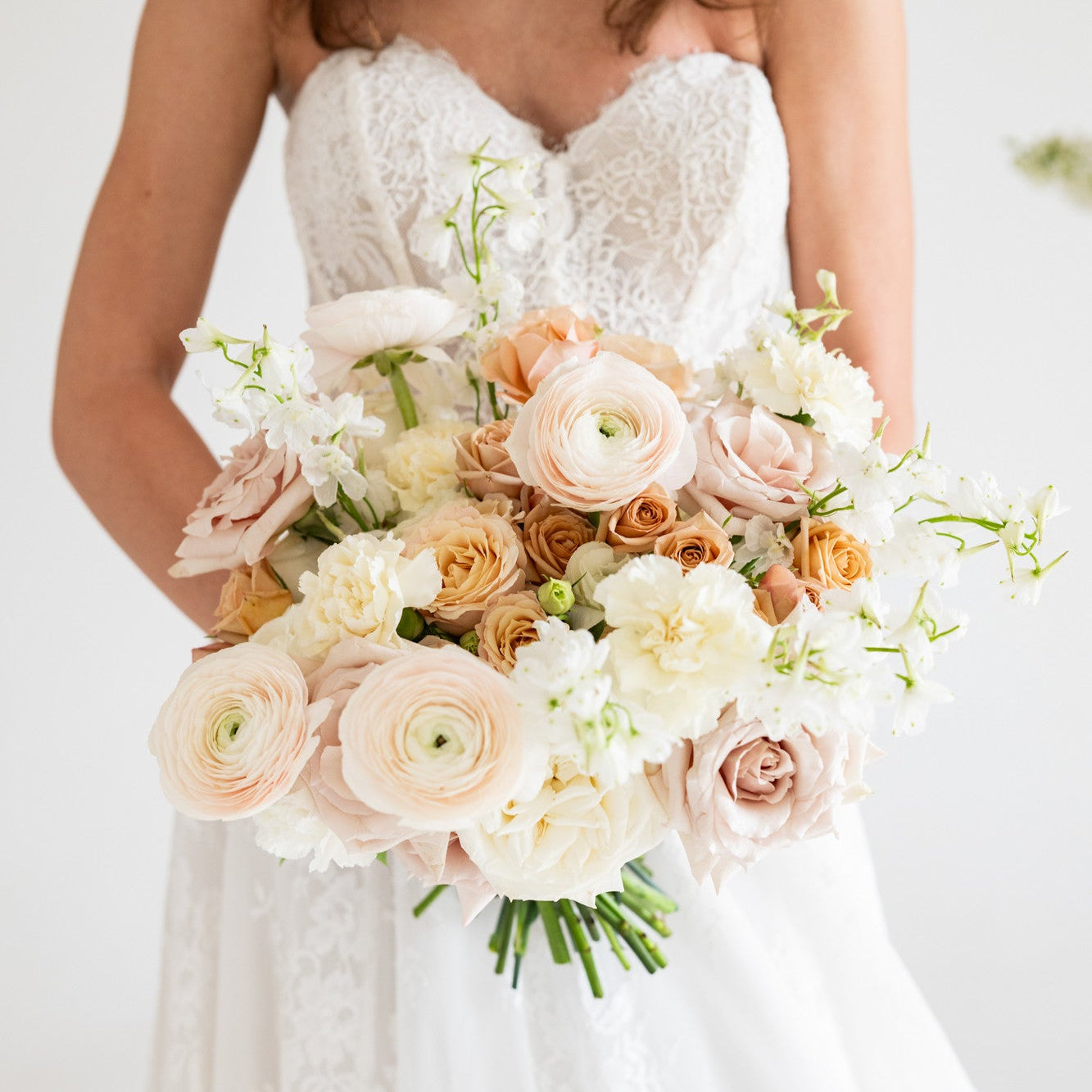A bride in a white lace wedding dress holding a large bridal bouquet of blush pink, peach, and white flowers against a bright white background.