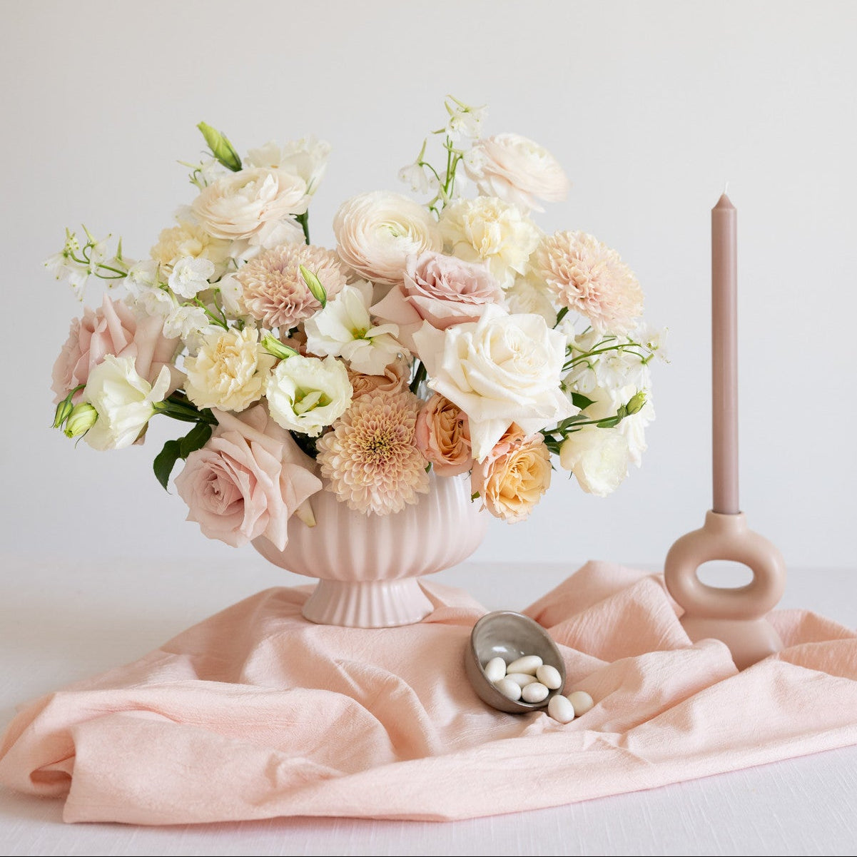 Centerpiece of flowers in neutral tones with candles and candy and a blush draped tablecloth. 