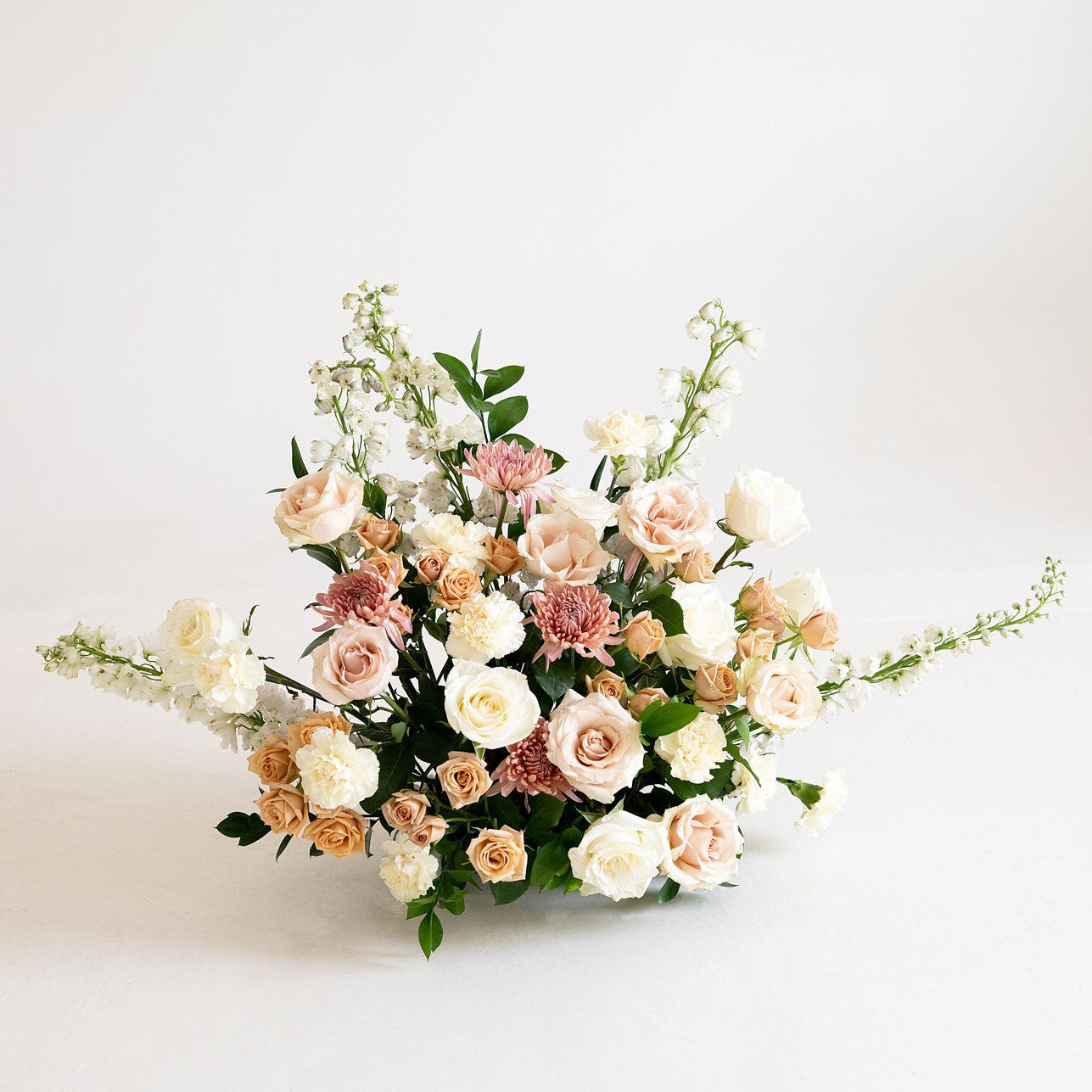 A delicate floral arrangement featuring blush pink, peach, and white roses with green foliage, displayed on a clean white background.