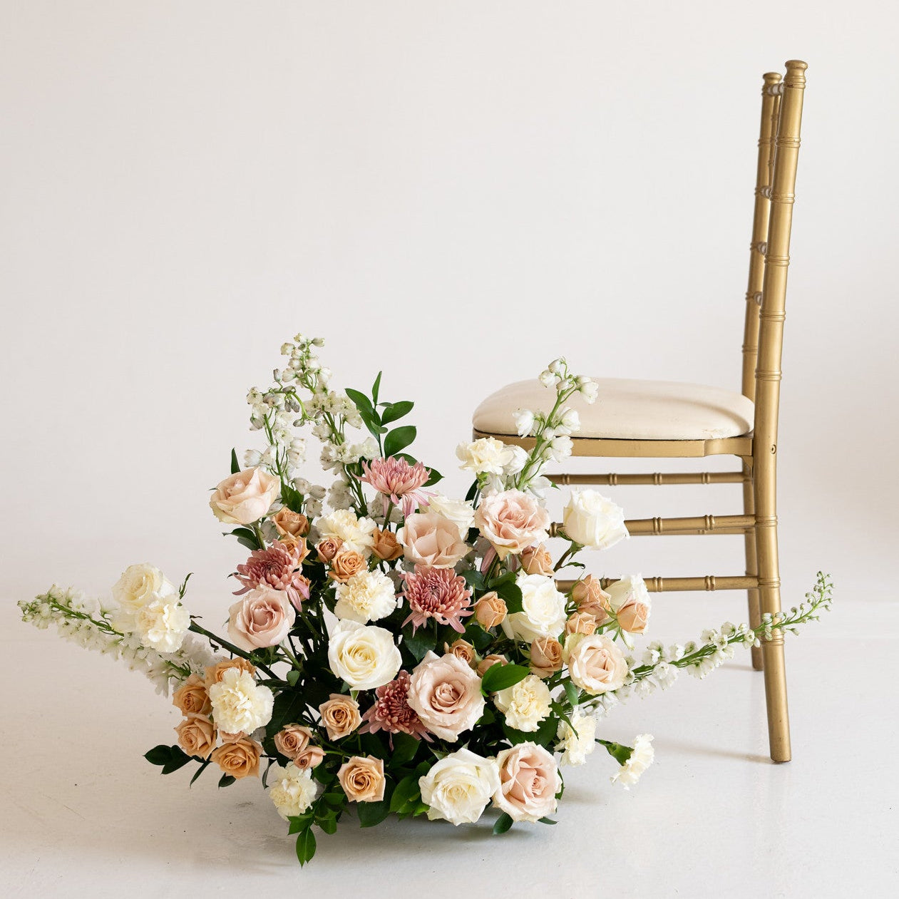 A large floral arrangement in blush pink, cream, and peach tones with green foliage, placed beside a gold Chiavari chair against a plain off-white background.