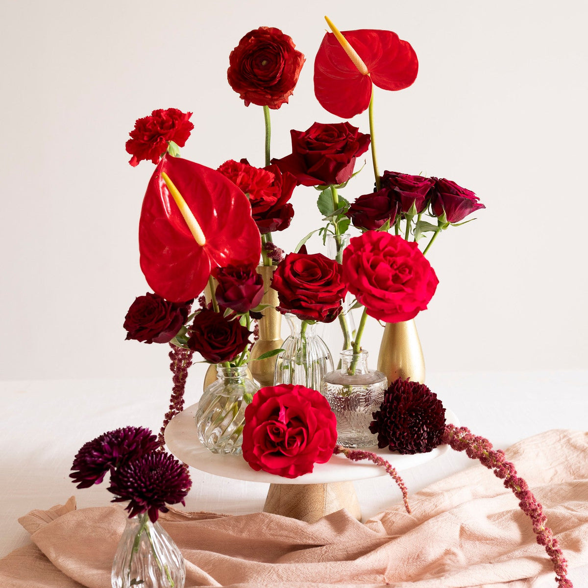 A styled floral arrangement of deep red roses, red anthuriums, and dark burgundy chrysanthemums in clear glass vases and a small gold vase, set on a draped blush pink fabric surface against a clean off-white background.