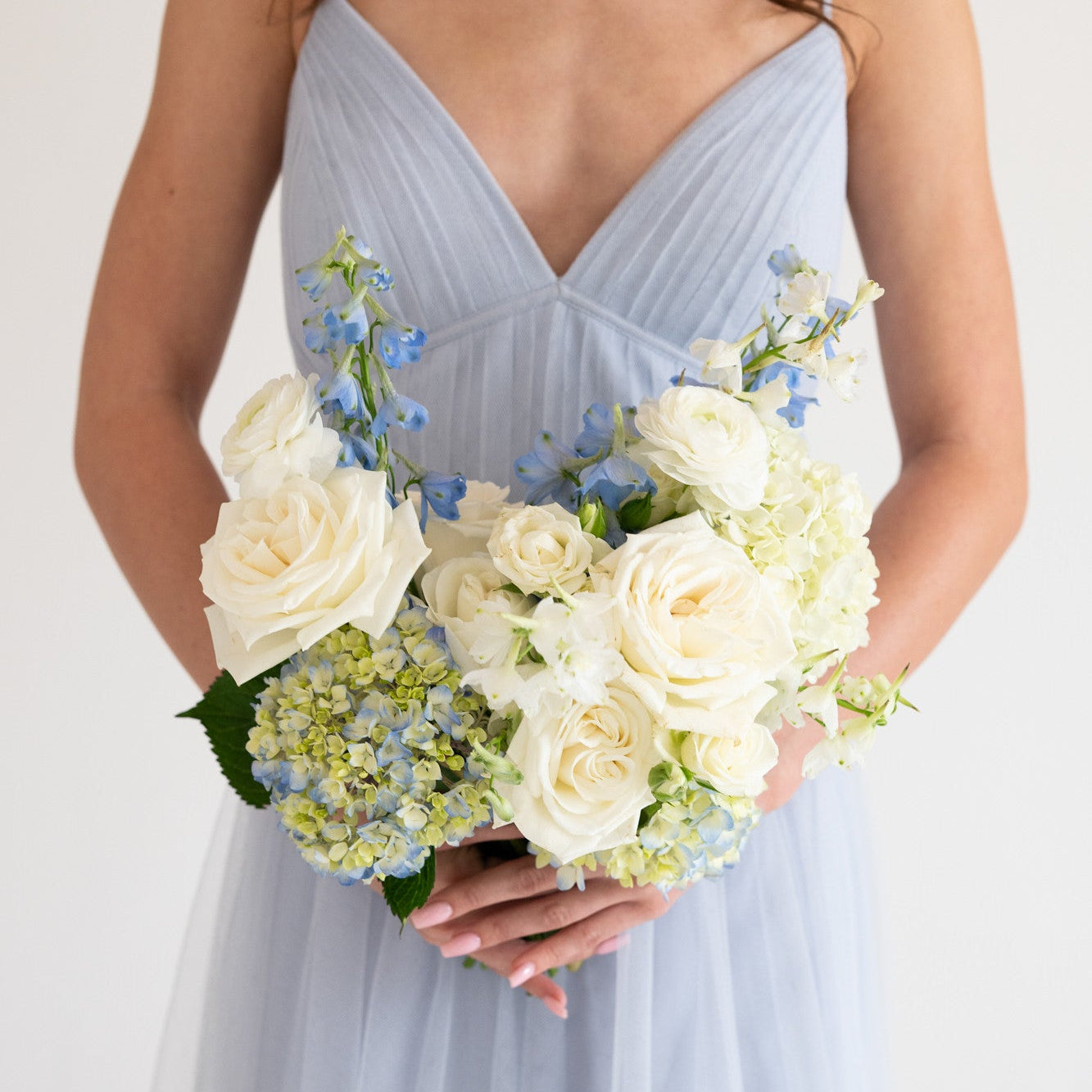A woman wearing a light blue pleated spaghetti-strap dress holds a bouquet of white roses, hydrangeas, and blue delphiniums against a plain white background.