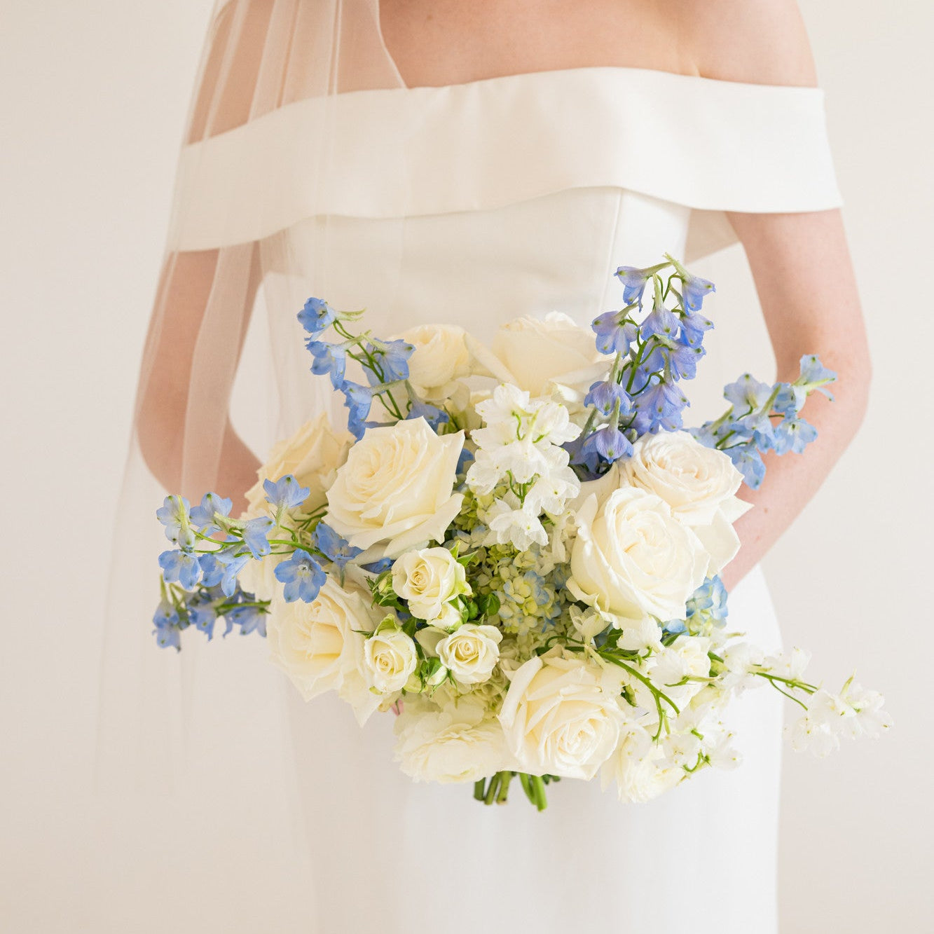 An off-the-shoulder white wedding dress with a sheer veil, held by a bride who is also holding a bouquet of white roses and blue delphiniums against a soft cream background.