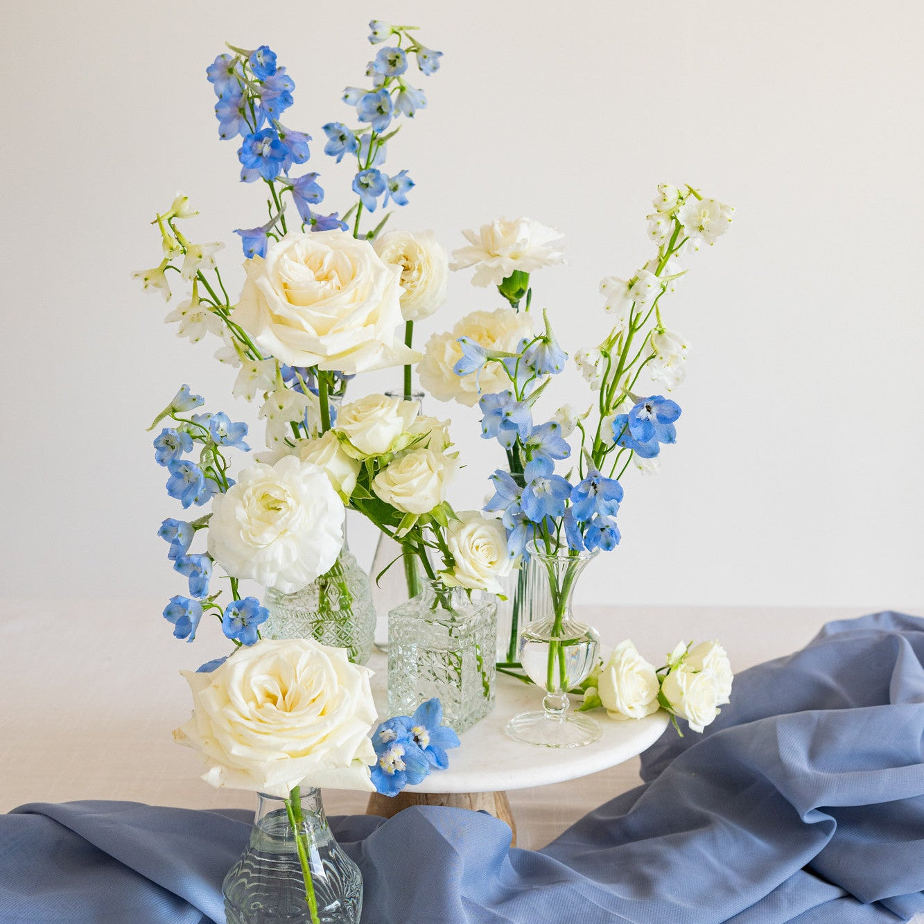 A floral bud vase assortment of white roses, blue delphiniums, and white carnations in clear glass vases, styled on a white table with a draped slate-blue fabric and a small round white stand against a light neutral background.