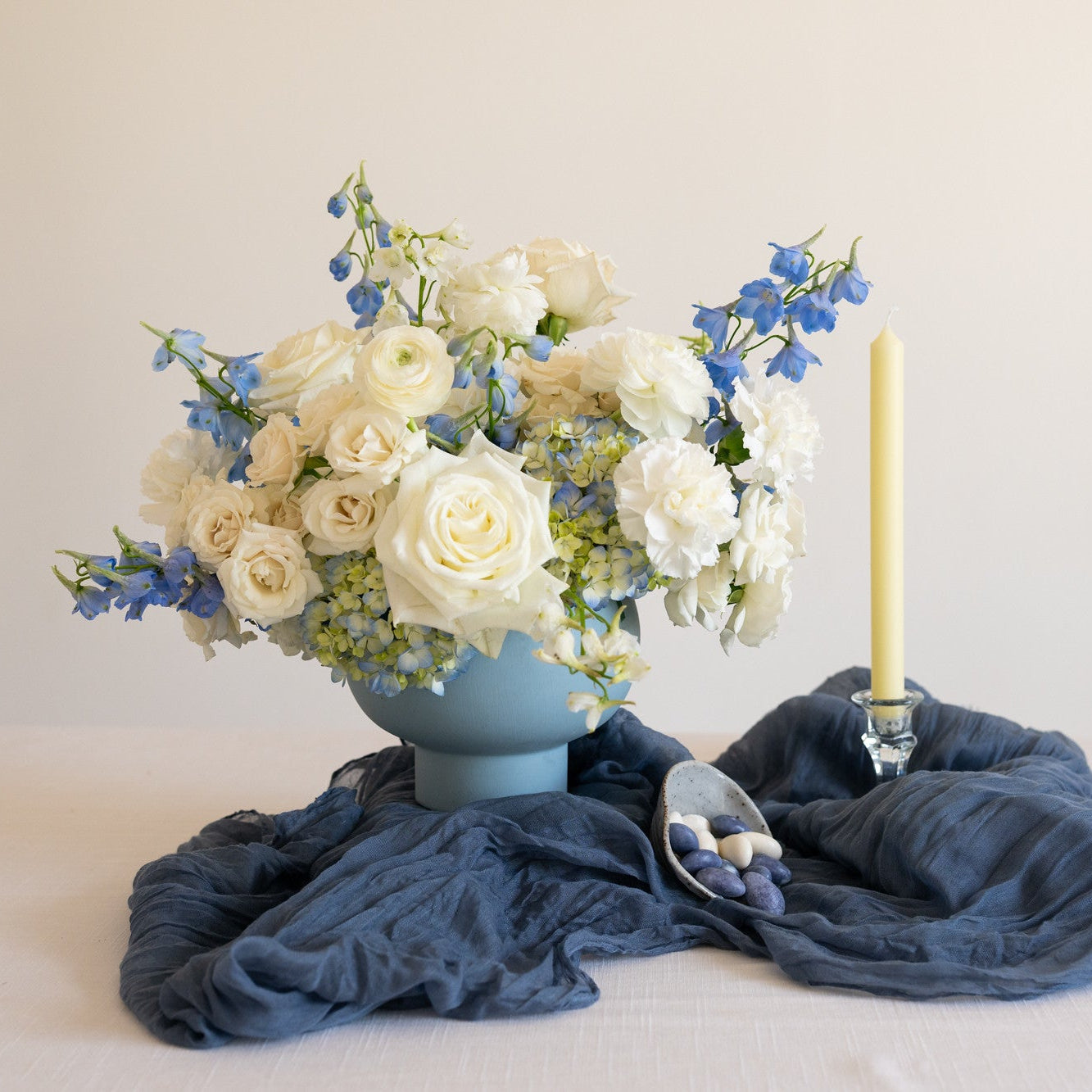 A floral arrangement of white roses, cream ranunculus, and blue delphiniums in a light blue vase, styled on a draped navy blue fabric with a single lit yellow taper candle and a small bowl of blueberries on a white table against a neutral beige background.